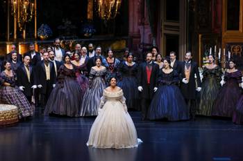 Μια ομάδα τραγουδιστών όπερας που φορούν κοστούμια της εποχής εμφανίζονται στη σκηνή, με περίτεχνα πολυέλαια και πολυτελή διακόσμηση στο παρασκήνιο.